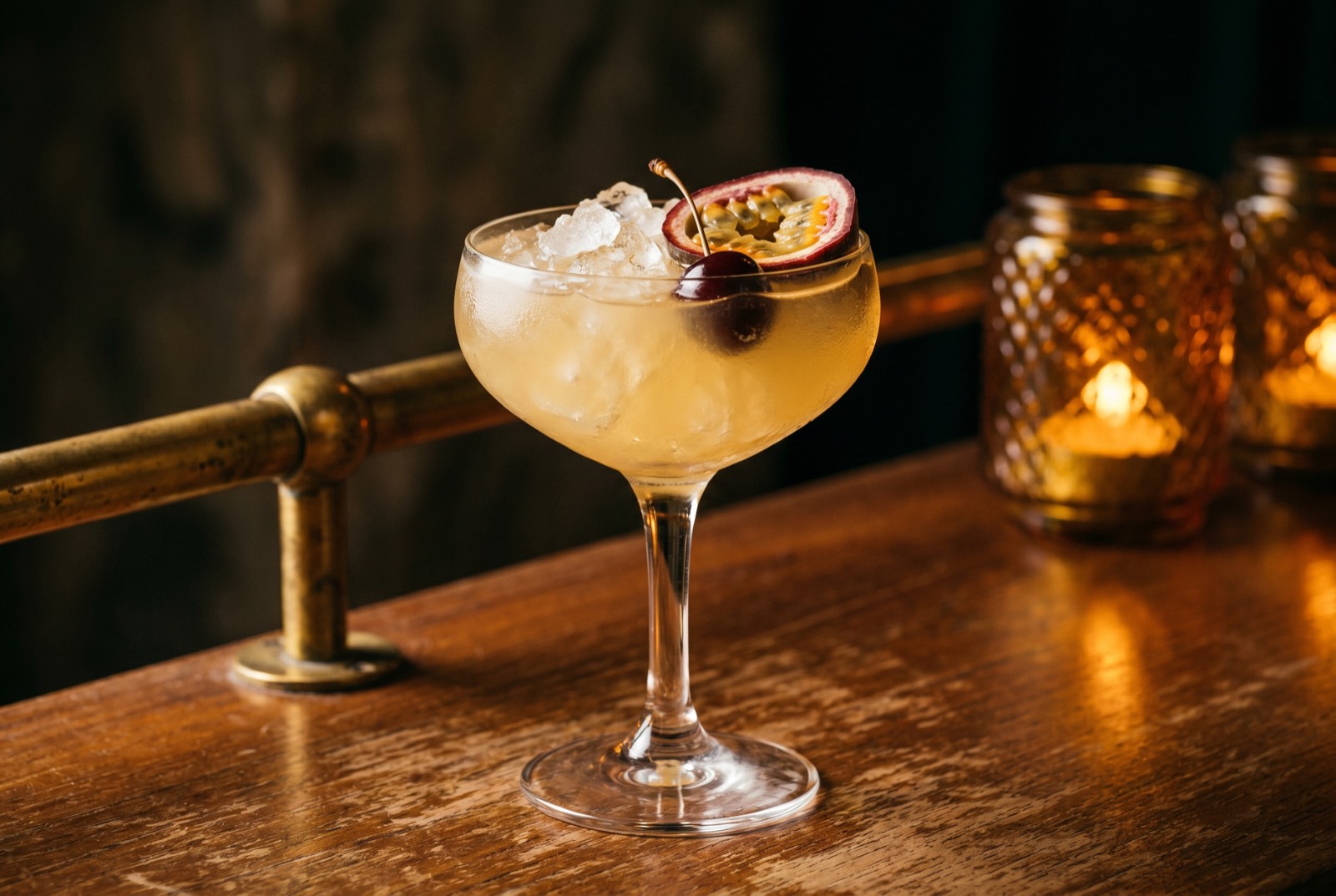Saturn cocktail, pale yellow-gold in a coupe glass over crushed ice with a cherry and passion fruit half, in the dim Forbidden Altar bar