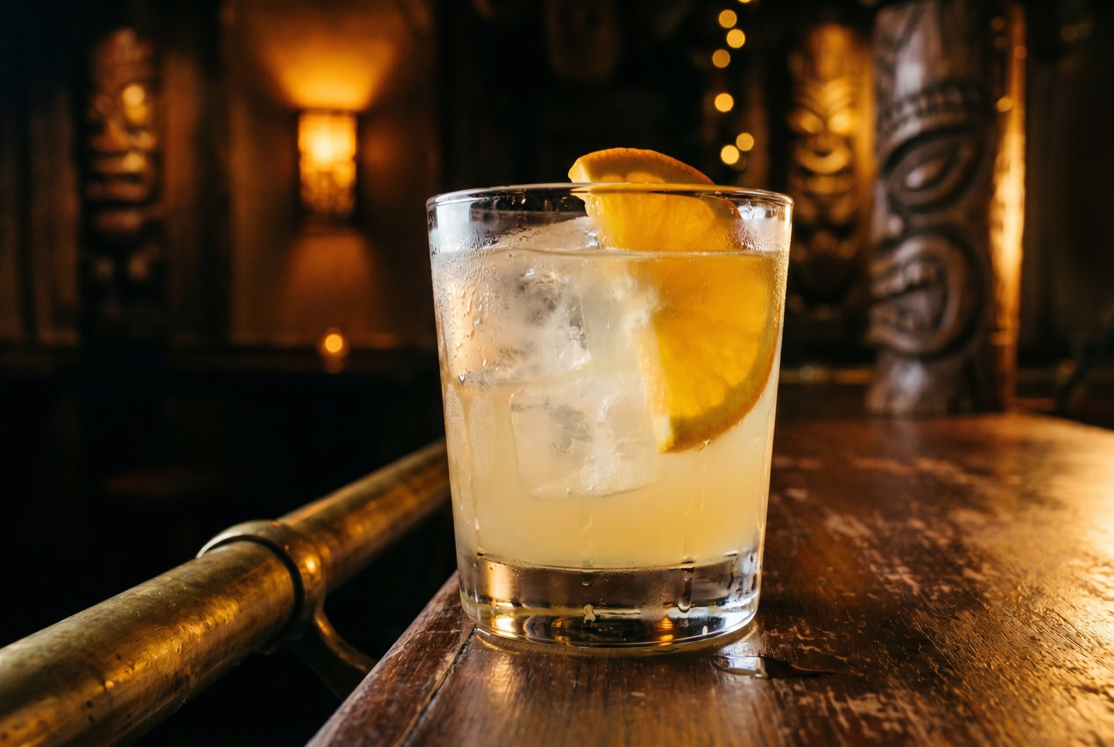 Under the Boardwalk cocktail, pale yellow in a rocks glass over ice with an orange slice, against the dark mask-lined Forbidden Altar bar